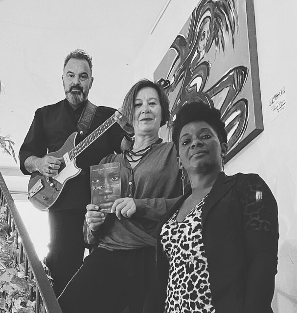 Swarz-Weiß Foto: Auf einer Treppe stehen von hinten oben nach vorne Sazan Azodi mit Gitarre, Anette Schmitt mit dem Buch "Kleiner Bruder" und die Sängerin Dianka Kuamé.