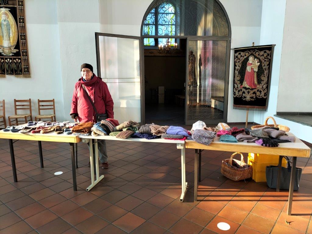 Auf einem langen Tisch liegen von links nach rechts gestickte Socken, Handschuhe, Schals und Mützen, die von einem Mitglied der Strickgruppe verteilt werden.
