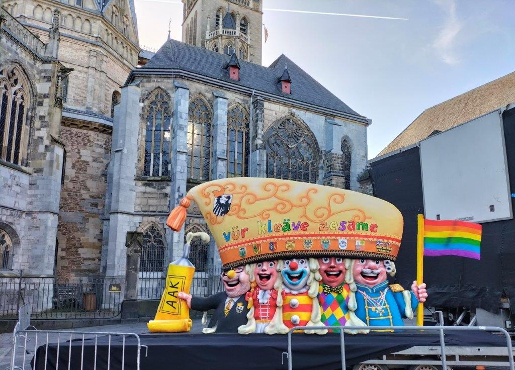Fünf Karnevalsfiguren unter einer Narrenkappe mit Aufschrift "vür kleäve zesame" halten eine Pride-Flagge und eine große Klebstofftube mit Aufschrift "AAK"