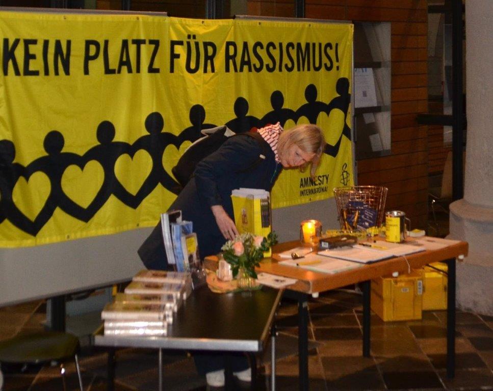 Amnesty Infostand und Buchtisch mit dem Buch "Durch die Wand" von der Seite, im Hintergrund ein Amnesty Banner mit Aufschrift "Kein Platz für Rassismus".