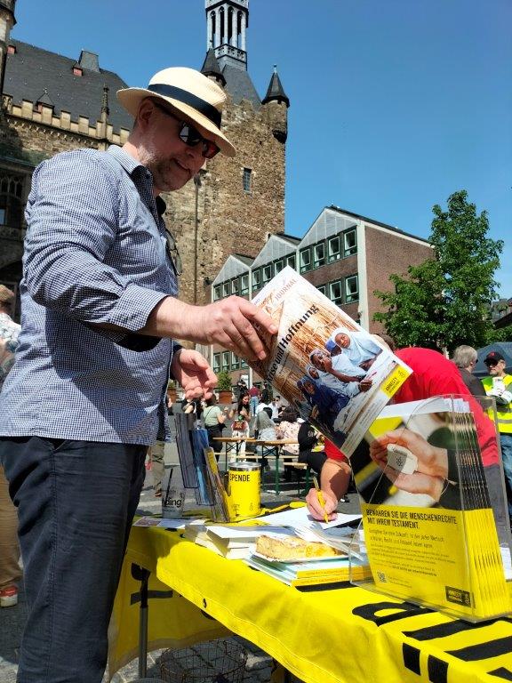 Mann nimmt Amnesty-Journal aus einem Zeitschriftenaufsteller am Amnesty-Infostand. Im Hintergrund sieht man einen Turm des Aachener Rathauses.