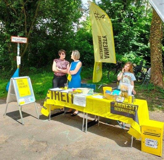 Menschen am Amnesty-Infotisch auf dem Aachener Weltfest 2024. Der Tisch ist mit einem gelben Amnesty-Banner gedeckt.