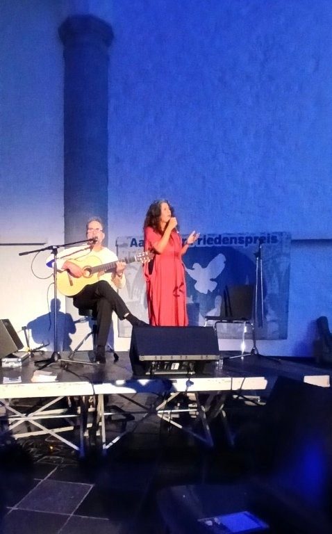 Eine Person spielt auf der Bühne Gitarre, eine Person in rotem Kleid singt, im Hintergrund ist ein Banner des Aachener Friedenspreis.