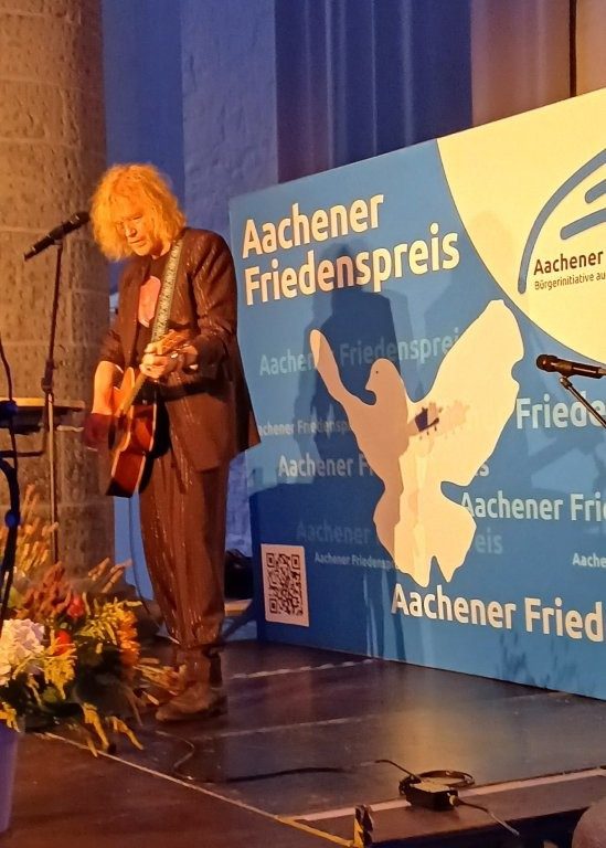 Eine Person spielt auf der Bühne Gitarre, im Hintergrund ist ein Banner des Aachener Friedenspreis.