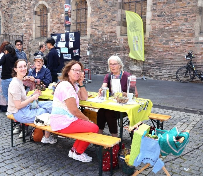 Mehrere Personen sitzen strickend an einer Bierzeltgarnitur, der Tisch ist mit einem Banner der Save-me Kampagne gedeckt. Auf einem kleinen daneben Wäscheständer sind verschiedene gestrickte Stücke ausgestellt.