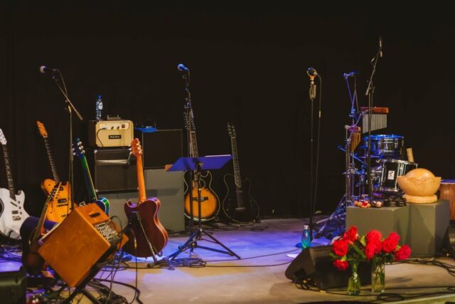 Eine Bühne in buntem Licht beleuchtet, keine Personen aber viele Musikinstrumente. Im Vordergrund zwei Vasen mit roten Rosen.