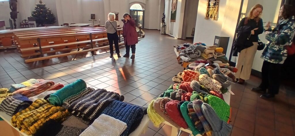 Einige Menschen stehen in einer Kirche und unterhalten sich, im Vordergrund sind drei Tische mit selbst gestrickten Dingen wie z.B. Schals und Mützen.