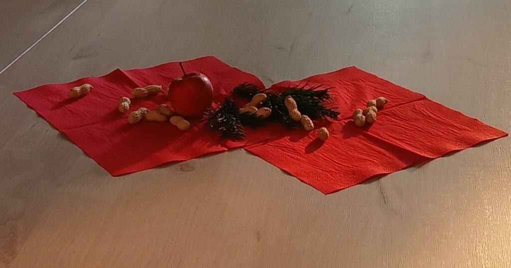 Zwei rote Servietten liegen auf einem Tisch, darauf Tannenzweige, ein Apfel und Erdnüsse.