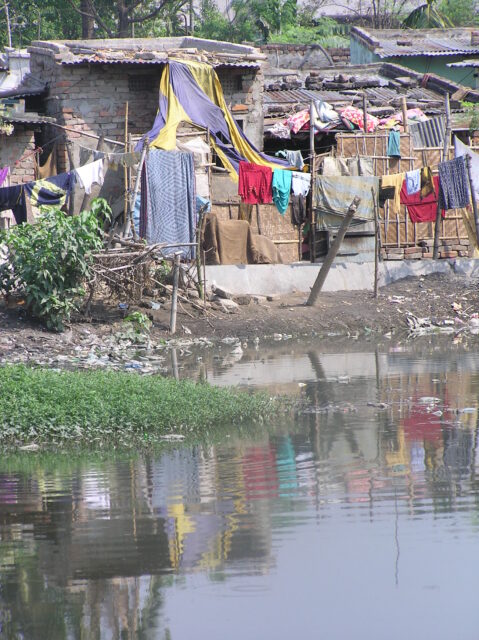Im Vordergrund ein Gewässer, dahinter schließt sich ein Gelände mit einfach gemauerten Hütten und Wellblechdächern an. Auf Wäscheleinen hängen verschiedene, bunte Wäschestücke zum trocknen.