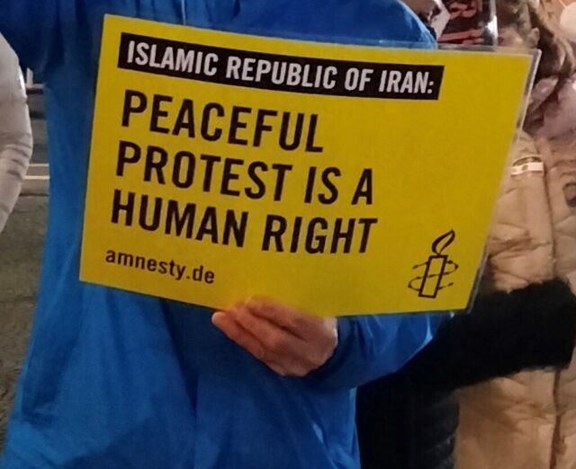 Person in blauer Regenjacke hält ein Amnestyplakat: "Islamic Republic of Iran: Peaceful protest is a human right".