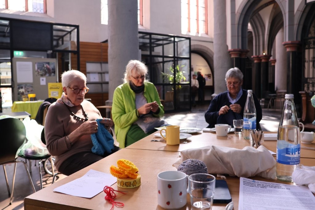 Mehrere Frauen sitzen mit Getränken und Tellern an einem Tisch und strickten in der Aachener Citykirche.