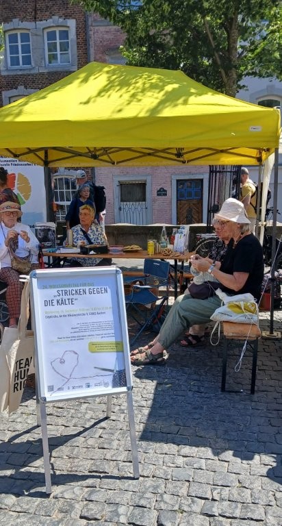 Mitglieder der Save-me Aachen Strickgruppe unter einem gelben Pavillon mit Infostand auf dem Katschhof in Aachen. Im Hintergrund sind weitere Infostände und Personen.