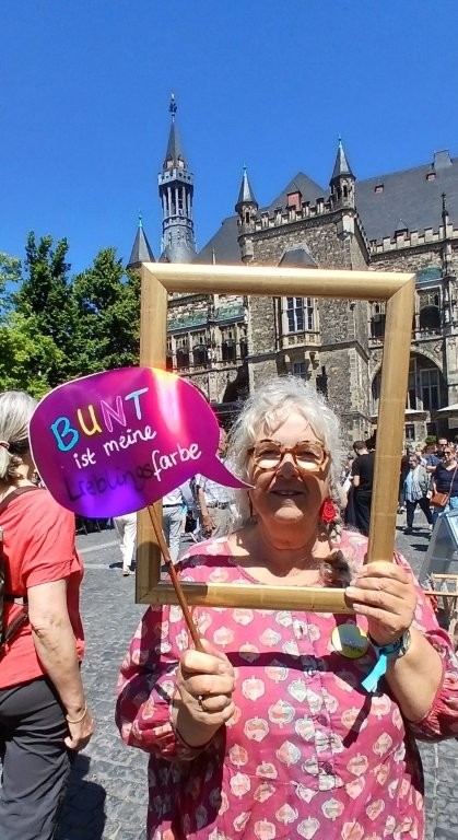 Foto einer Frau, die einen Bilderrahmen und eine pinke Sprechblase mit den Worten "BUNT ist meine Lieblingsfarbe" hält. Im Hintergrund das historische Aachener Rathaus.