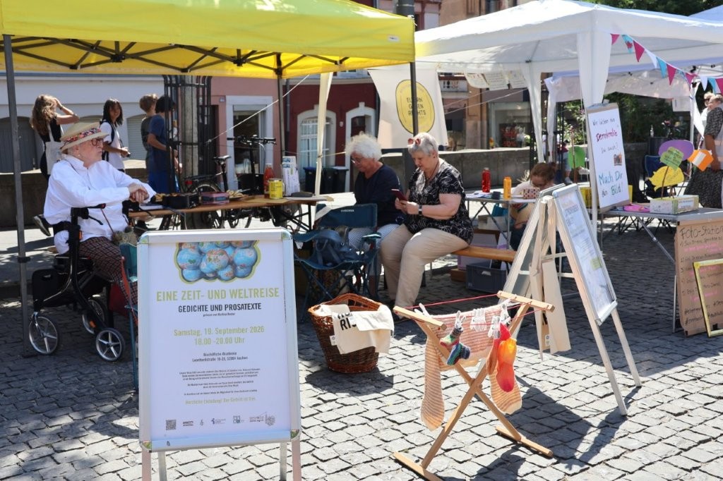 Mitglieder der Save-me Aachen Strickgruppe unter einem gelben Pavillon mit Infostand auf dem Katschhof in Aachen. Im Hintergrund sind weitere Infostände und Personen.