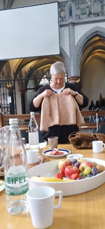 Eine Frau hält eine gestrickte Kinderjacke vor hinter einem gedeckten Tisch in der Aachener Citykirche.